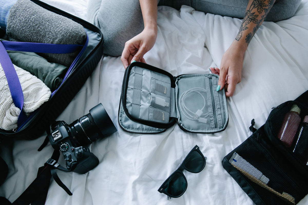 A person organizing a travel packing cube on a bed next to a suitcase, camera, and sunglasses