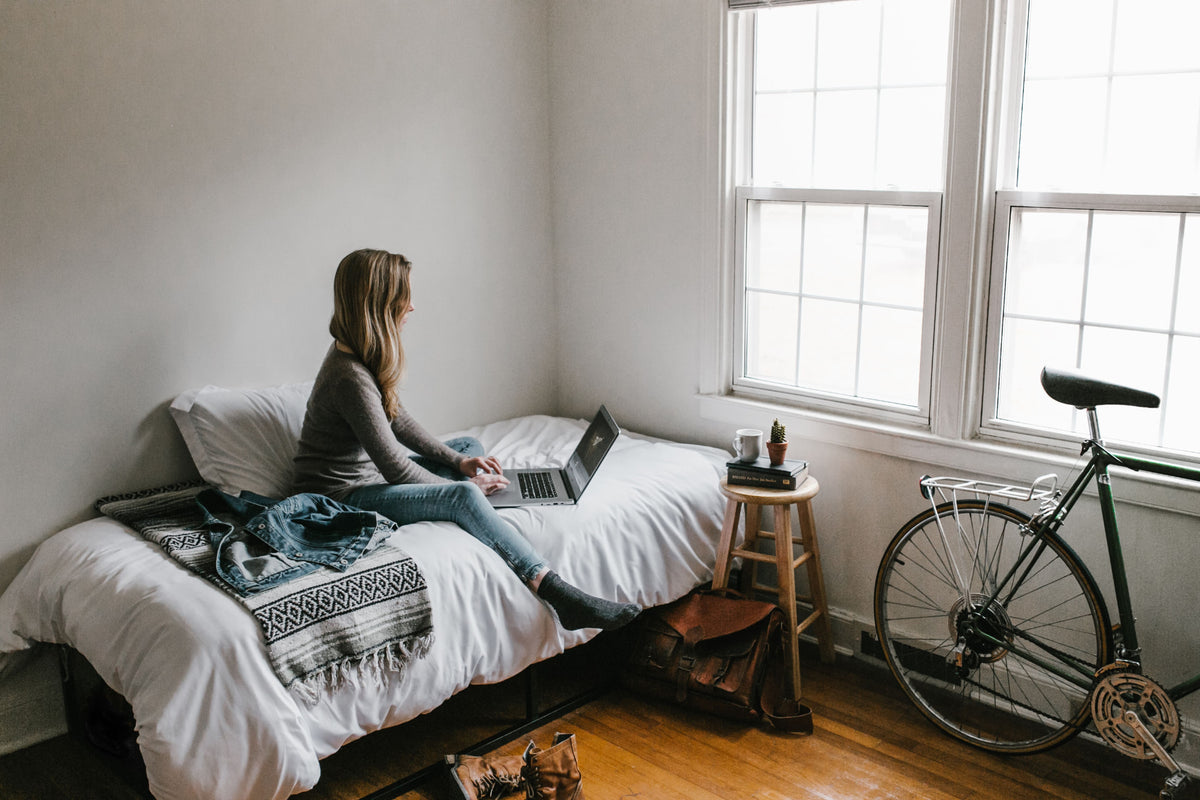 A young woman sitting on a dorm bed working on a laptop in a bright room with a bicycle by the window