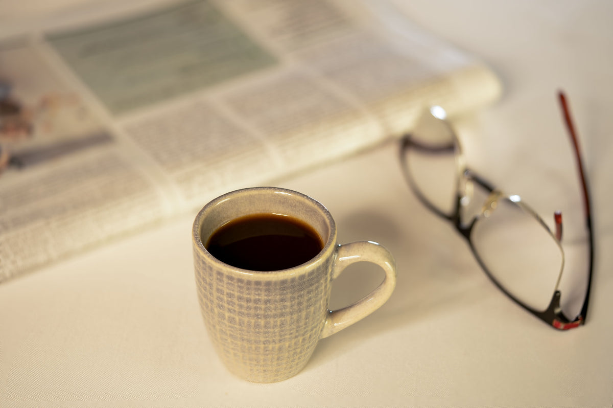 A ceramic mug of black coffee on a table beside reading glasses and a newspaper