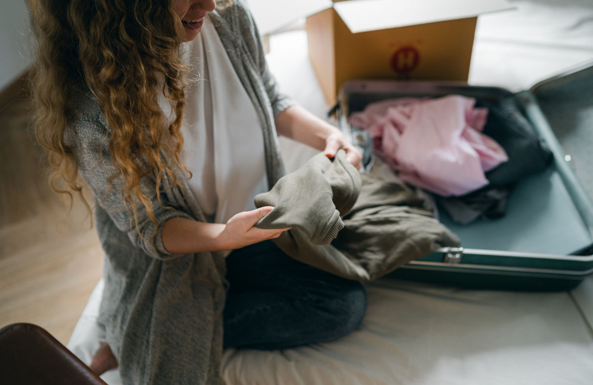 A young woman folding clothes into an open suitcase on a bed while packing for college move-in day