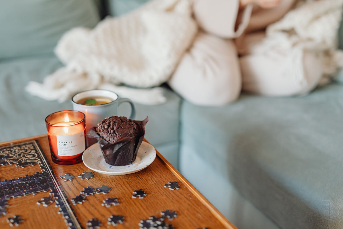 A lit candle, a mug of tea, and a muffin on a wooden tray beside a cozy sofa