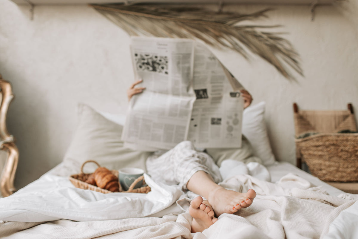 A person reading the newspaper while sitting up in bed with croissants and a mug of tea on a wicker tray