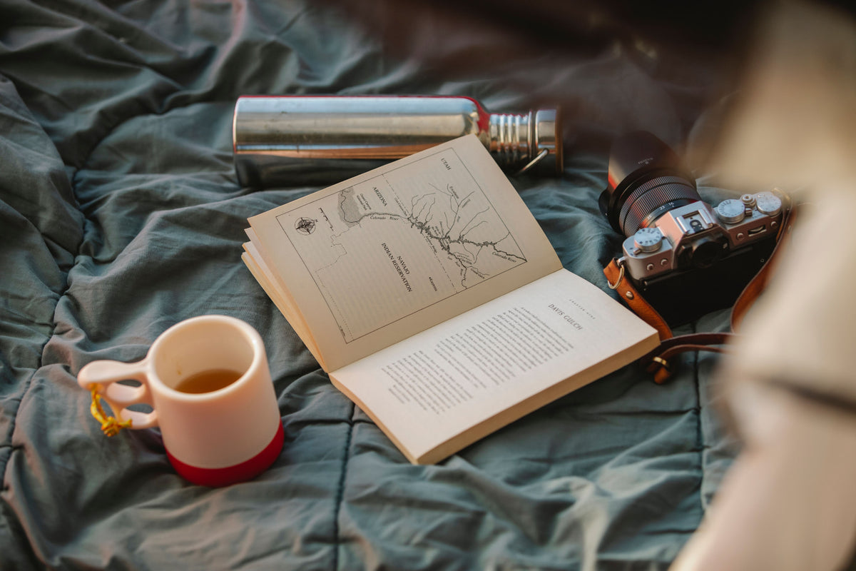 A travel wellness flat lay with an open book, a mug of tea, a water bottle, and a camera on a soft blanket
