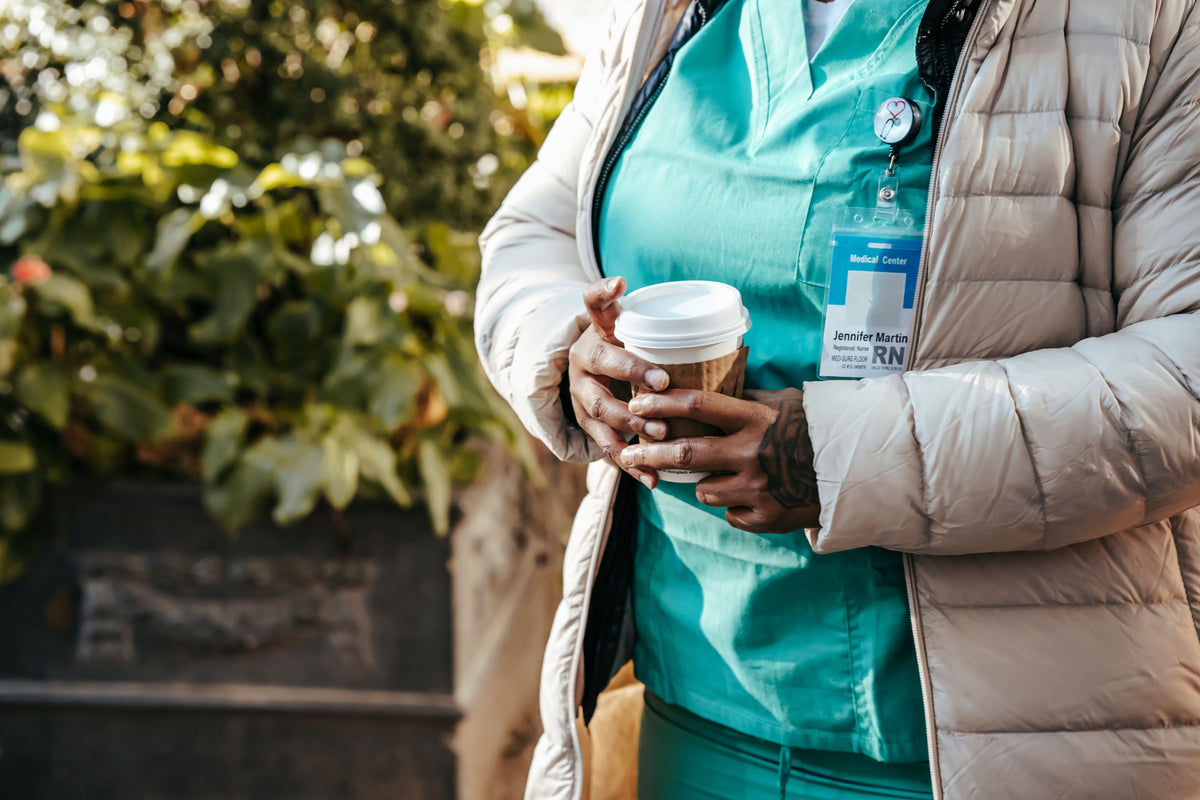 A travel nurse in teal scrubs and a tan puffer jacket holding a coffee cup outdoors